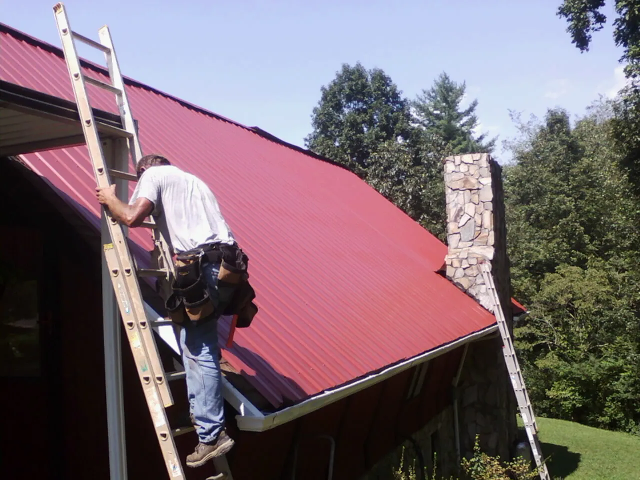 Roofer on a ladder beside a red metal roof and stone chimney — Soffit & Fascia Repair by Roofing Brothers in Porter Ranch